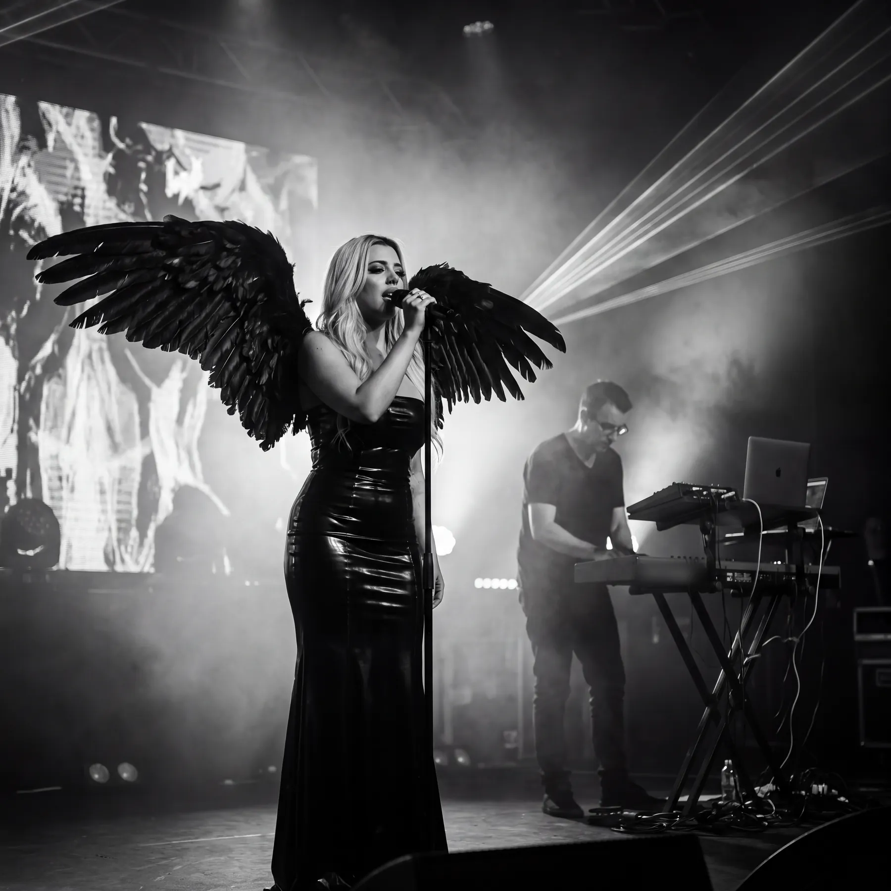Kat singing as Regina on stage wearing large black feathered wings, the producer at keyboards behind her in haze and laser light